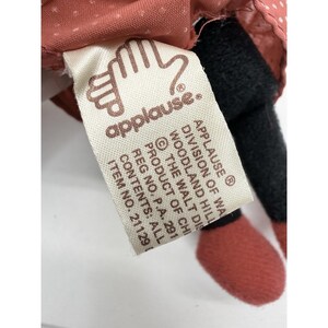 May include: A close-up of a doll with a tag that reads "applause." The doll has a pink dress with white polka dots, black pants, and red shoes. The tag includes the text "APPLAUSE DIVISION OF WA WOODLAND HILLS THE WALT DIS PRODUCT OF CHI REG NO. P.A. 291 CONTENTS: ALL ITEM NO. 21129."