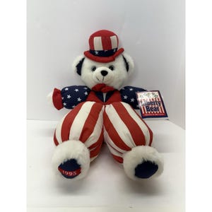 May include: A plush Liberty Bear doll, dressed in patriotic attire. The bear wears a red, white, and blue top hat, a bow tie, and striped pants. The arms are adorned with blue fabric and white stars. The year "1995" is embroidered on the foot.