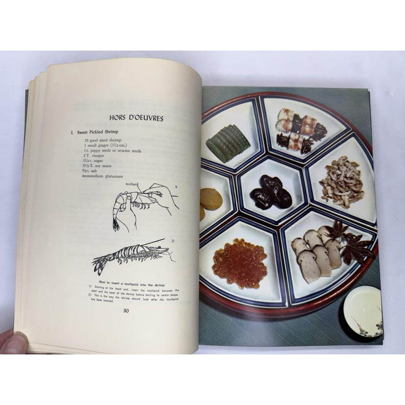 May include: An open cookbook page with the title "HORS D'OEUVRES" and a diagram of a shrimp. A serving tray with various appetizers, including caviar, mushrooms, and pickled vegetables, is on the right page.
