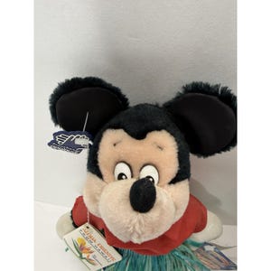 Puede incluir: Mu&ntilde;eco de peluche de Mickey Mouse con una camisa roja y una falda de hierba azul y verde. El mu&ntilde;eco tiene orejas negras y una etiqueta que dice "Aloha Friends From Hawaii". El mu&ntilde;eco es un objeto de colecci&oacute;n de Disney.