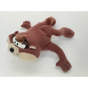 May include: A plush toy of the Tasmanian Devil, a cartoon character. The toy is primarily brown with a tan face and white eyes. The toy is lying on its stomach with its arms and legs outstretched.