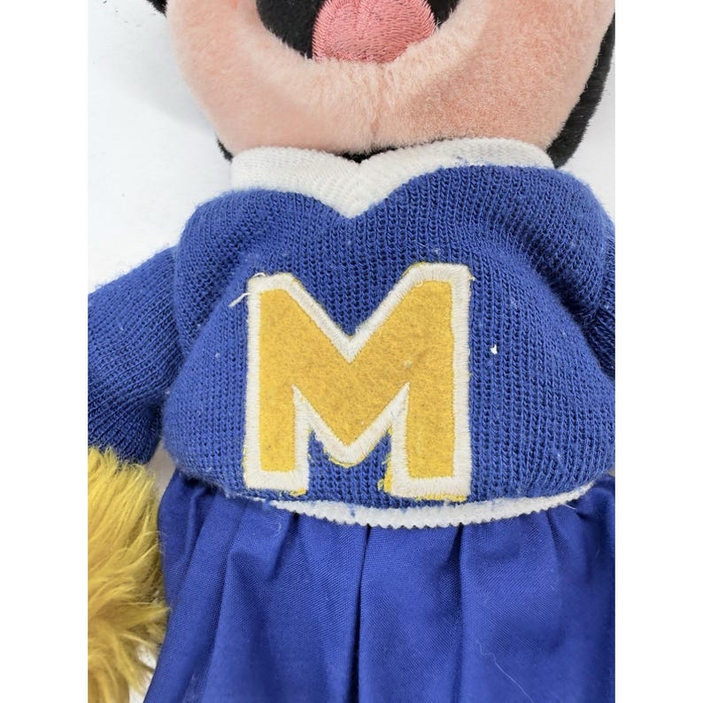 May include: A plush doll wearing a blue sweater with a large yellow "M" and a blue pleated skirt. The doll has a pink face, black eyes, and a black and white head. A yellow pom-pom is visible on the left side.