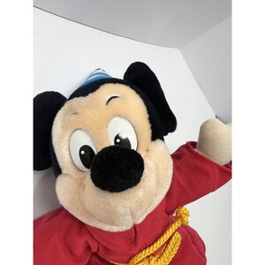 May include: A plush Mickey Mouse toy wearing a red robe and a blue and white hat. The toy has a yellow rope tied around its waist.