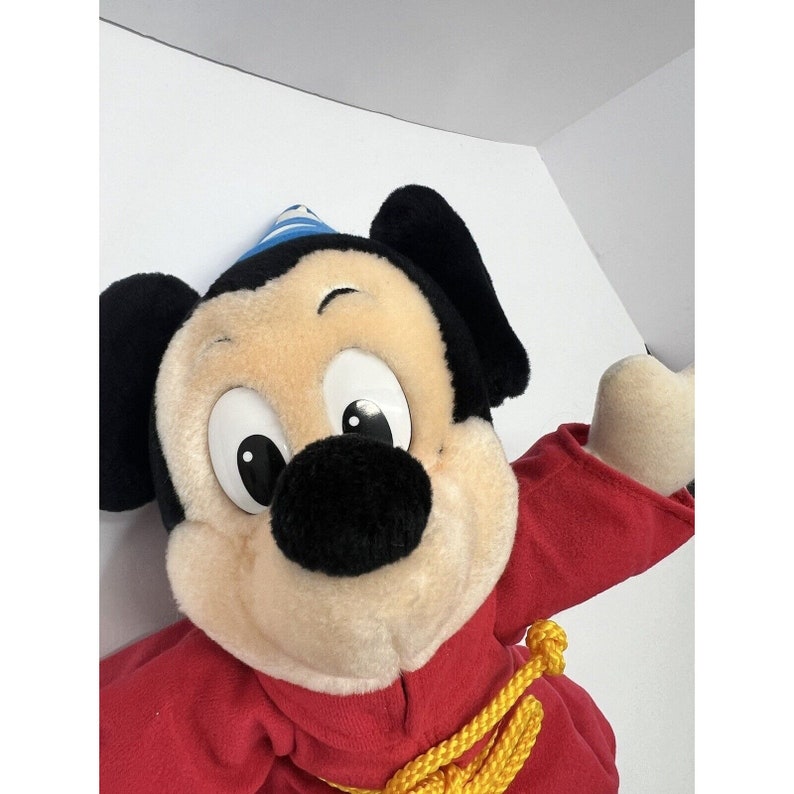 May include: A plush Mickey Mouse toy wearing a red robe and a blue and white hat. The toy has a yellow rope tied around its waist.
