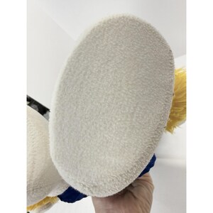 May include: Close-up of a plush toy's foot, featuring a soft, off-white sole. The foot is part of a larger stuffed animal, with visible yellow hair and blue accents. The toy is held against a white background.
