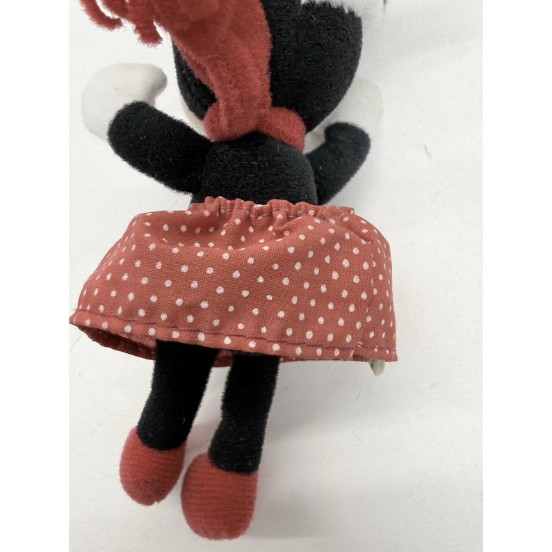 May include: A plush doll of a cartoon character, wearing a red polka-dot skirt, black top, and red scarf. The doll has black legs and red shoes. White gloves and a white hat complete the outfit.