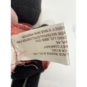 May include: A close-up of a tag from a Disney X'Mas Mini Prepack plush toy. The tag is white with black text, listing the manufacturer's information, including the company name, address, and material composition.