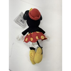 Puede incluir: Un peluche de Minnie Mouse, con una falda roja con lunares amarillos, un sombrero rojo y zapatos amarillos. El peluche negro tiene guantes blancos y una etiqueta adjunta.