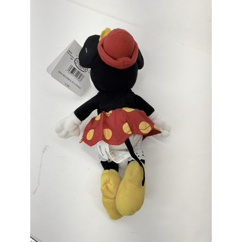 Puede incluir: Un peluche de Minnie Mouse, con una falda roja con lunares amarillos, un sombrero rojo y zapatos amarillos. El peluche negro tiene guantes blancos y una etiqueta adjunta.