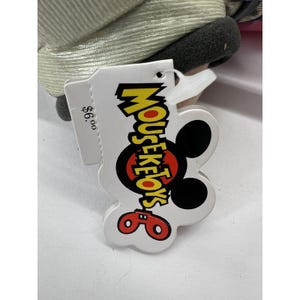 May include: A white tag with a black Mickey Mouse head and the text "Mouseketoys" in yellow letters. The tag also has a red price tag that says "$6.00".
