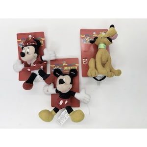 May include: Three Disney plush toys: Minnie Mouse in a red polka-dot dress, Mickey Mouse in red shorts, and Pluto the dog. Each toy is attached to a red cardboard backing with the "Mickey Cuddly" logo.