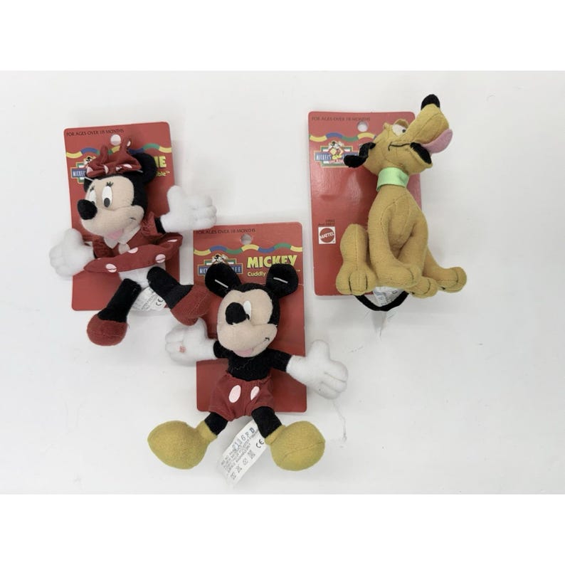 Puede incluir: Tres peluches de Disney: Minnie Mouse con un vestido de lunares rojos, Mickey Mouse con pantalones cortos rojos y Pluto el perro. Cada juguete est&aacute; unido a un respaldo de cart&oacute;n rojo con el logo "Mickey Cuddly".