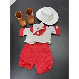 May include: A doll outfit consisting of a white cowboy hat with a yellow star emblem, a pair of brown plastic boots, a red and white collared shirt with red buttons, and matching red pants. The set is designed for a doll and features a western theme.