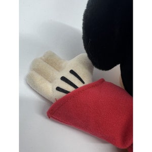 May include: Close-up of a plush Mickey Mouse toy's hand. The hand is white with black stitching for the fingers and a red sleeve.