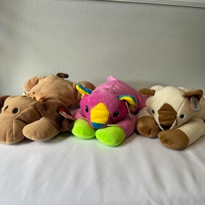 May include: Four plush animal toys in a row. The toys include a brown dog, a tan bear, a pink and yellow creature, and a cream-colored bear with brown accents. Each toy has a tag attached.