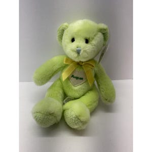 May include: A lime green plush teddy bear with a yellow ribbon bow and a heart-shaped tag that reads "August". The bear has black eyes and a light blue nose. It is sitting with its arms and legs outstretched.