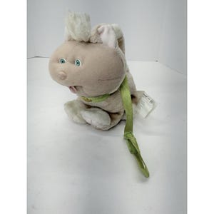 May include: A plush bunny toy in beige with light green accents. The bunny has blue eyes, a pink tongue, and a green ribbon around its neck. The toy has a tag attached to it.