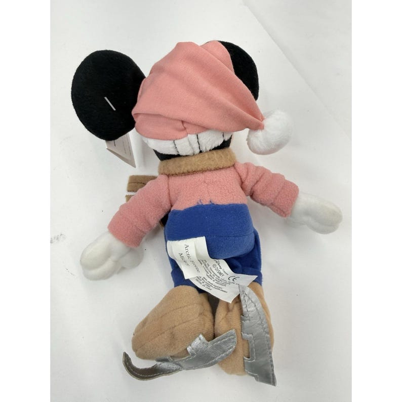 May include: A plush Mickey Mouse doll wearing a pink winter hat with a white pom-pom, a pink and blue outfit, and brown and silver ice skates. The doll has black ears and white gloves. A tag is attached to the doll.