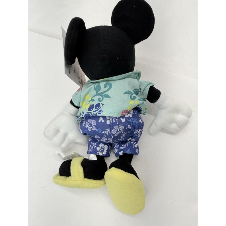 May include: A Mickey Mouse plush toy wearing a Hawaiian shirt and shorts. The shirt is light blue with floral patterns, and the shorts are blue with white flowers. The toy has black ears, a black head, white gloves, and yellow shoes.