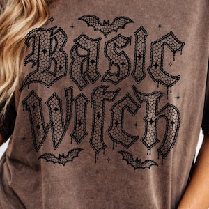 May include: Brown t-shirt featuring the text "Basic Witch" in a gothic font, with bat illustrations. The shirt has a vintage, worn appearance. The model is wearing blue denim shorts.