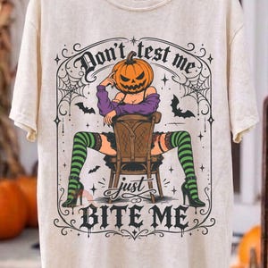 Könnte beinhalten: Ein cremefarbenes T-Shirt mit einer Grafik einer Figur mit Kürbiskopf, die auf einem Stuhl sitzt und ein violettes Oberteil sowie grün gestreifte Strümpfe trägt. Der Text lautet "Don't test me just BITE ME", umgeben von Spinnennetzen und Fledermäusen.