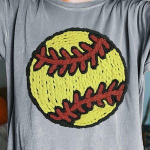 May include: Gray t-shirt featuring a large, embroidered softball design. The softball is yellow with red stitching and a black outline. The shirt has a relaxed fit and short sleeves, suitable for casual wear.