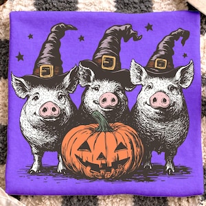 May include: A purple t-shirt featuring three pigs wearing witch hats and an orange jack-o'-lantern. The pigs are smiling, and stars are scattered around the design. A fun Halloween-themed graphic.