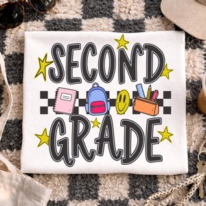 Puede incluir: Camiseta blanca con las palabras "SECOND GRADE" en negro y amarillo, con gráficos escolares. Incluye un cuaderno, una mochila, una carita sonriente y un estuche. Gafas de sol y gorra beige visibles.