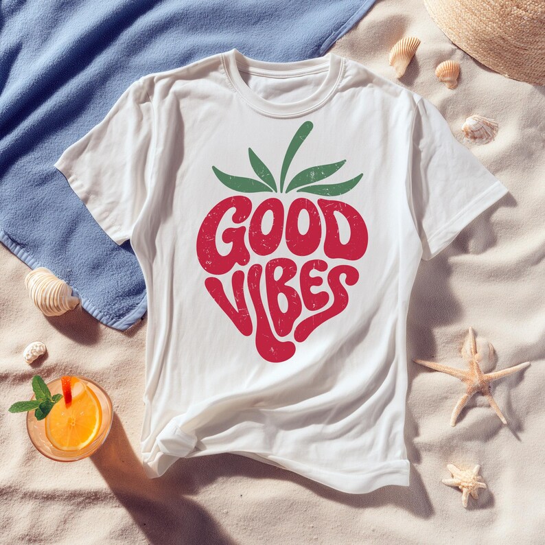 May include: White t-shirt with a red strawberry design that reads "GOOD VIBES" in a retro font. The strawberry has green leaves. The shirt is on a sandy surface with a blue towel, seashells, a straw hat, and a drink.