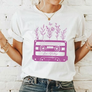 May include: White t-shirt with a pink vintage cassette tape design. The cassette tape has flowers and a butterfly growing out of it. The text "Love Vintage" is printed on the cassette tape.