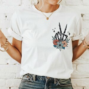 May include: White t-shirt featuring a black skeleton hand making a peace sign surrounded by colourful flowers.