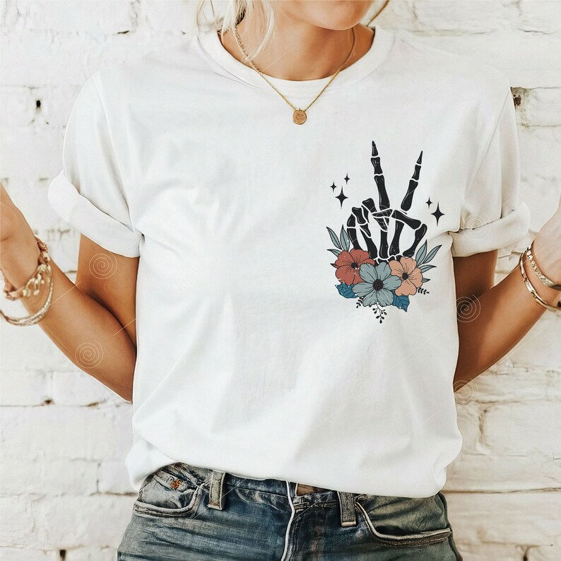May include: White t-shirt featuring a black skeleton hand making a peace sign surrounded by colourful flowers.
