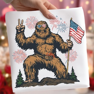 May include: A white square fabric featuring a brown Bigfoot character wearing sunglasses with an American flag design. The Bigfoot is holding an American flag and giving a peace sign, with red and blue fireworks in the background.