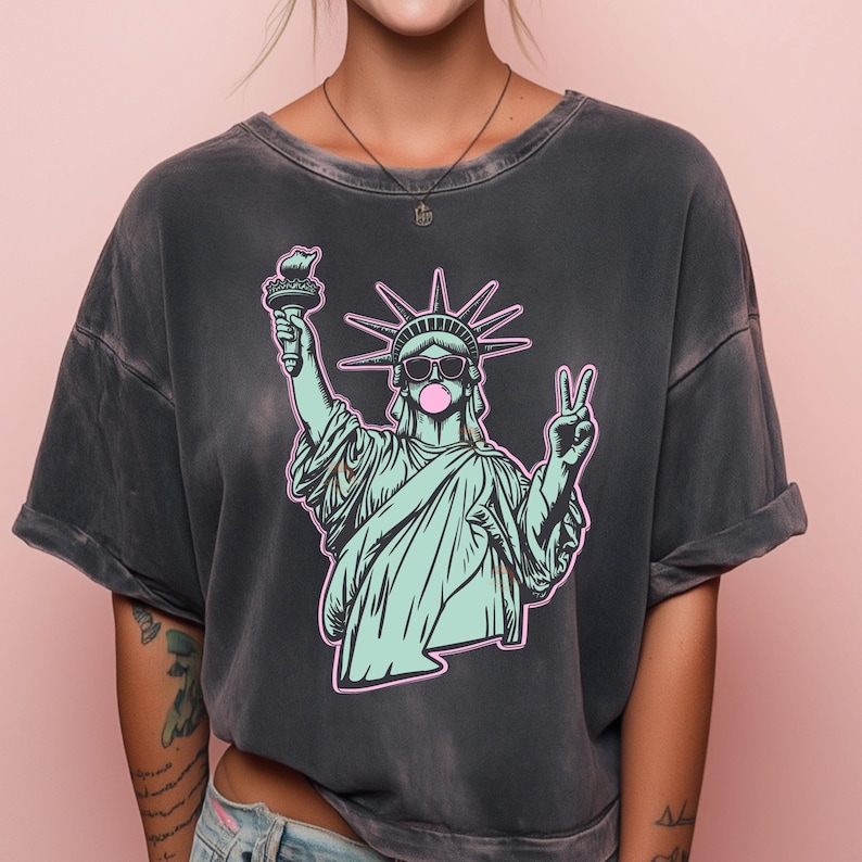 May include: Dark gray t-shirt featuring a graphic of the Statue of Liberty wearing sunglasses and blowing a bubblegum bubble. The Statue of Liberty is holding a torch and making a peace sign. The graphic is outlined in pink.