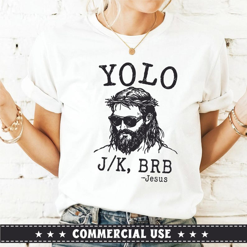 May include: White t-shirt with a black graphic of a person wearing sunglasses and a crown of thorns. The text "YOLO" is above the graphic, and "J/K, BRB -Jesus" is below. The shirt is worn by a person.