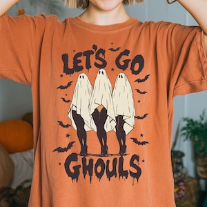 Może przedstawiać: Pomarańczowa koszulka z napisem "LET'S GO GHOULS" w ciemnobrązowym kolorze. Koszulka przedstawia grafikę trzech duchów z czarnymi nogami i nietoperzami. Zabawna koszulka na Halloween.