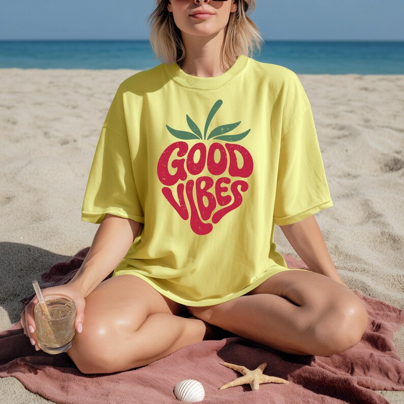 May include: A yellow t-shirt with a red strawberry design that says "GOOD VIBES". The shirt is worn on a beach with a drink in hand. The design is a retro style, perfect for summer.
