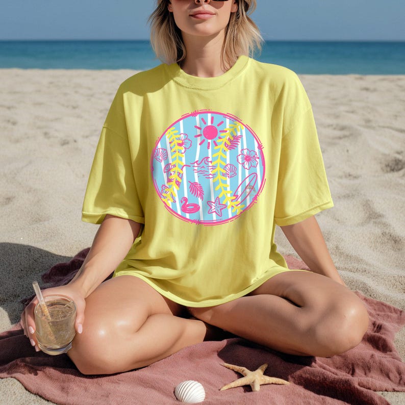May include: Yellow oversized t-shirt featuring a circular graphic with a beach theme. The graphic includes a pink sun, blue waves, and various beach-related icons like a surfboard, flowers, and a flamingo. The shirt is worn on a beach setting.