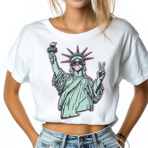 May include: White crop top featuring a graphic of the Statue of Liberty wearing sunglasses and blowing a bubblegum bubble. The Statue of Liberty is holding a torch and making a peace sign with her other hand. The graphic is outlined in pink.