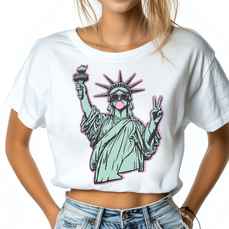 May include: White crop top featuring a graphic of the Statue of Liberty wearing sunglasses and blowing a bubblegum bubble. The Statue of Liberty is holding a torch and making a peace sign with her other hand. The graphic is outlined in pink.