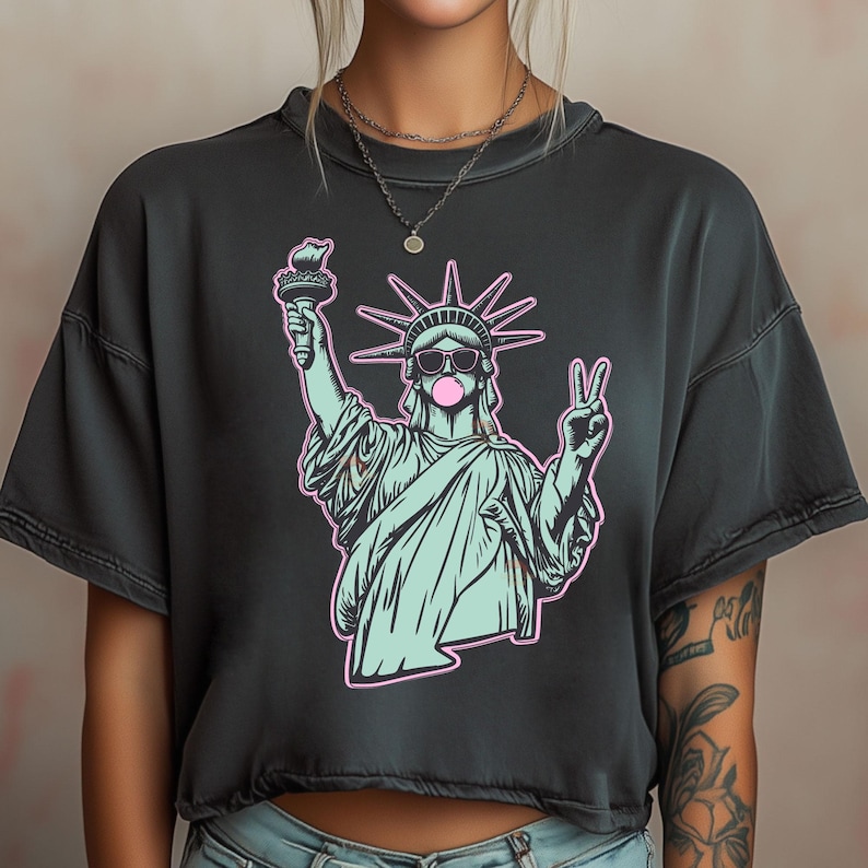 May include: Dark gray t-shirt featuring a graphic of the Statue of Liberty wearing sunglasses and blowing a bubble. The statue is in a light blue color with pink outlines, holding a torch and making a peace sign.