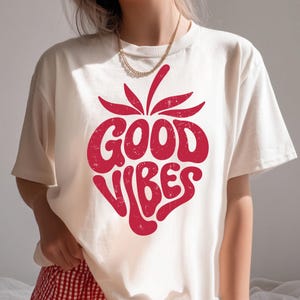 May include: Cream-colored t-shirt with a red strawberry graphic. The strawberry design features the words "GOOD VIBES" in a bold, retro font. The shirt is made of a soft material and has short sleeves.