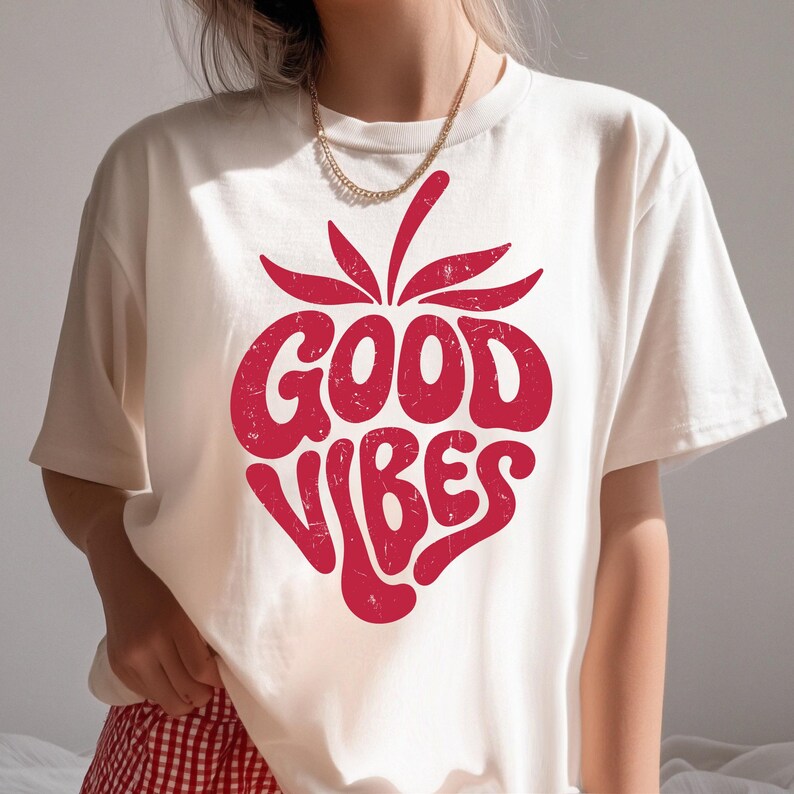 May include: Cream-colored t-shirt with a red strawberry graphic. The strawberry design features the words "GOOD VIBES" in a bold, retro font. The shirt is made of a soft material and has short sleeves.