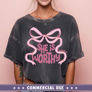 May include: A dark gray t-shirt with a pink bow and the text "She is worthy" and "Proverbs 31" printed on it.