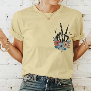 May include: A yellow t-shirt featuring a black skeleton hand making a peace sign, surrounded by pink, blue and white flowers.