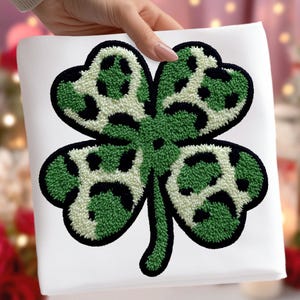 May include: A four-leaf clover patch with a green and white leopard print design. The clover is outlined in black and has a textured, embroidered appearance. The patch is on a white background.