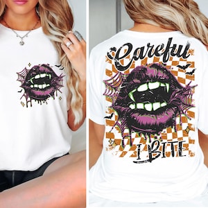 May include: White t-shirt featuring a graphic of purple lips with fangs, surrounded by a spiderweb design. The back of the shirt has the text "Careful I Bite" over a checkered orange and white background, with bat and star accents.