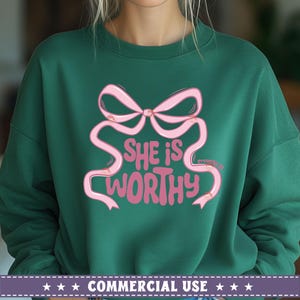 May include: A green sweatshirt with a pink bow and the text "She is worthy" in a pink, cursive font. The text "Proverbs 31" is in a smaller font below the bow.