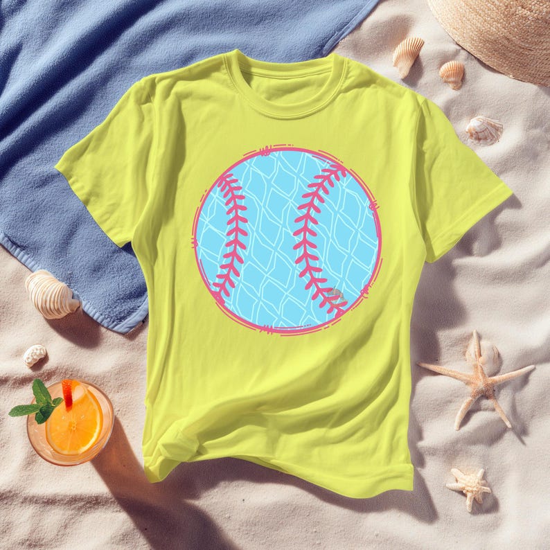 May include: A yellow t-shirt featuring a large baseball graphic. The baseball design is light blue with pink stitching and a pink outline. The shirt is on a sandy surface with a blue towel, seashells, a straw hat, and a drink.