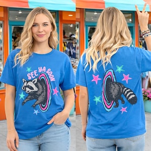 May include: Blue t-shirt featuring a raccoon graphic emerging from a portal with the text "See Ya Soon." The design includes pink, blue, and green accents with star shapes. The back of the shirt also features the raccoon and star graphics.